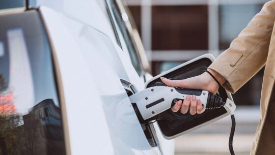 Woman in a beige coat plugging in an electric vehicle to charge.