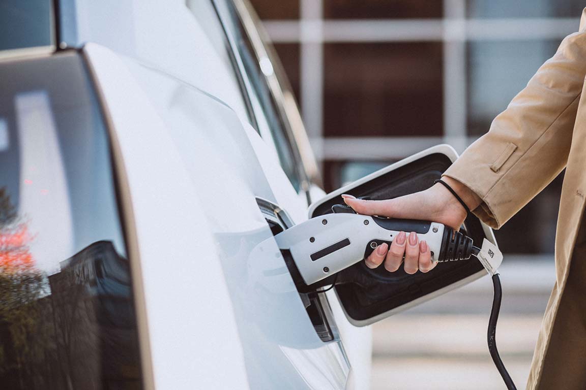 Woman in a beige coat plugging in an electric vehicle to charge.